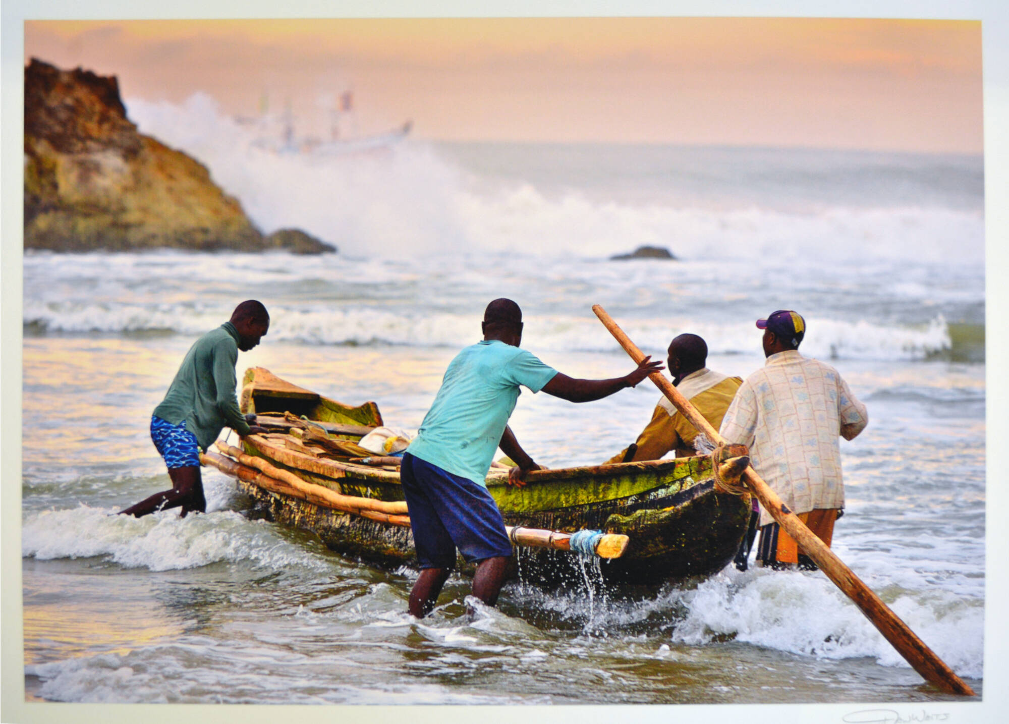 Four men launching a boat with long oar, waves crashing on the rocks in the background. Taken in Ga District, Ghana.